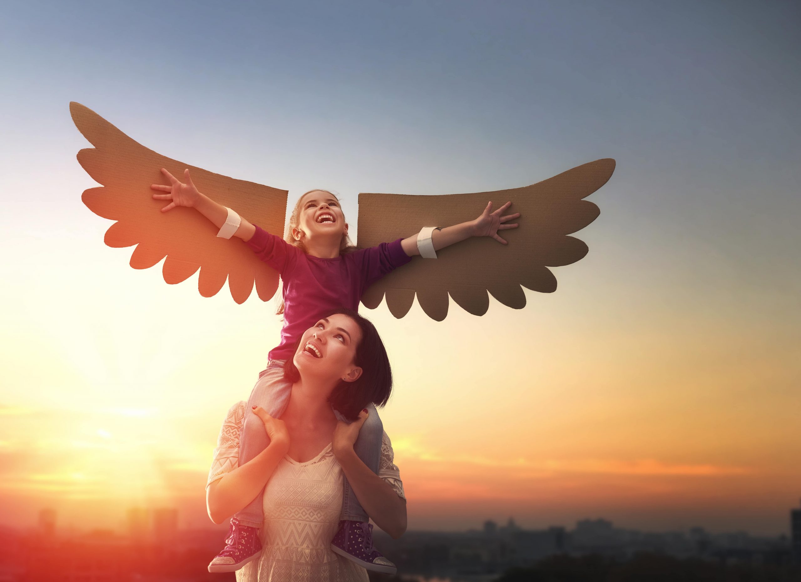 nysa-therapy-banner A woman has a toddler on her shoulders. The toddler has cardboard wings on her arms and is pretending to fly. Sunset in the background.